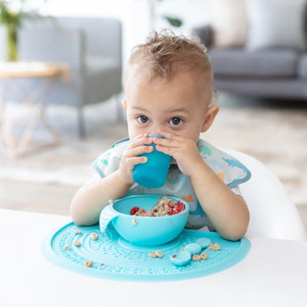 A toddler sips from a blue Bumkins silicone starter cup at the table, wearing a patterned bib with a matching bowl and food mat.