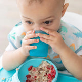 In a bib, the baby drinks from a Bumkins Silicone Starter Cup: Blue, with oatmeal and berries in front of them.