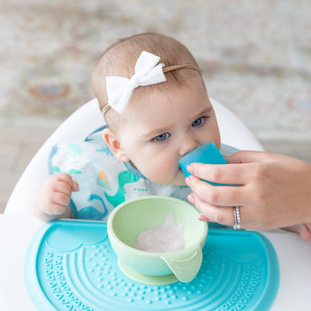 A baby in a bib and bow sits in a high chair learning to drink from the Bumkins Silicone Starter Cup: Blue, guided by an adult hand.