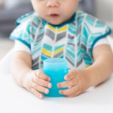 A baby in a patterned bib holds a Bumkins Silicone Starter Cup: Blue, seated happily at a white table.