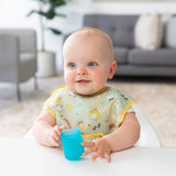 A baby uses a Bumkins Silicone Starter Cup: Blue while sitting at a table in a bright room with plants and a gray couch in the background.