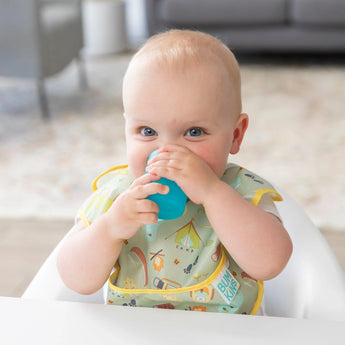 Baby in a high chair joyfully sipping from a Bumkins Silicone Starter Cup: Blue, wearing a patterned bib.