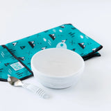 Bumkins Silicone Stretch Lid for Grip Bowl with spoon on a folded teal and black cloth on a white surface.