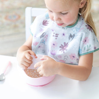 Young girl in a floral bib eating cereal from a bowl secured with Bumkins Silicone Stretch Lid for Grip Bowl.