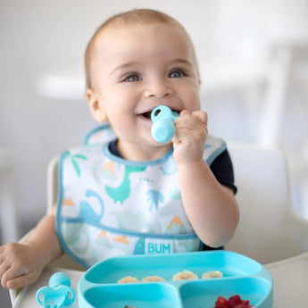 A baby in a dinosaur bib explores weaning with Bumkins Silicone Chewtensils®: Blue, eating from a divided plate at a high chair.