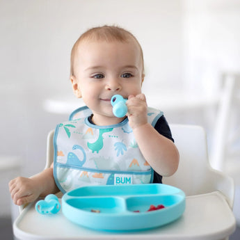 A baby in a high chair wears a Bumkins Starter Bib with dinosaur prints, holding a small blue object and enjoying snacks from a matching blue silicone divided plate on the tray, both featuring waterproof fabric for easy cleaning.