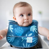 Baby wearing a Bumkins Blue Tropic Starter Bib, in a high chair looking sideways; its waterproof and machine washable.