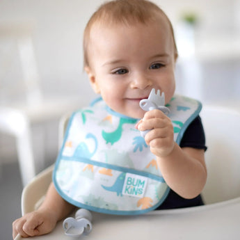 A baby in Bumkins Starter Bib from the Dinosaurs & Blue Tropic 2 Pack, holds a gray spoon in a high chair. Its machine washable!.