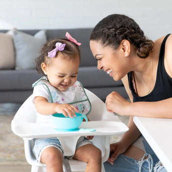 A woman and a toddler with a bow smile at each other over Bumkins Starter Bib 2 Pack: Floral & Lace, toddler happily gripping bowl.