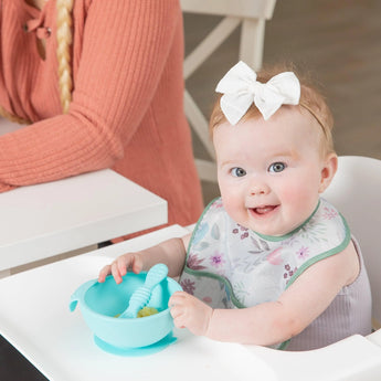 A smiling baby with a white bow holds a blue bowl in her high chair as Bumkins Starter Bibs make mealtime easy to clean.