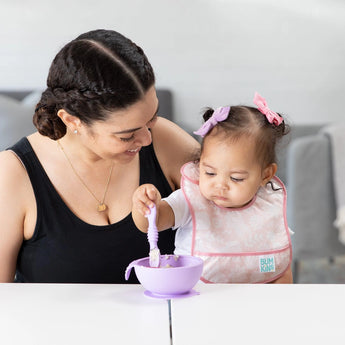 Mother smiles as baby, in a Bumkins Starter Bib Floral & Lace, reaches for a purple bowl at the table.