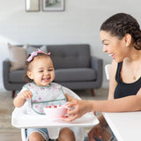 A smiling mom feeds her happy toddler in a Bumkins Starter Bib 2 Pack: Floral & Lace, which makes cleaning food spills easy.