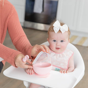 Smiling baby in high chair with white bow wears Bumkins Starter Bib. Easy-clean waterproof fabric. Kitchen in background.