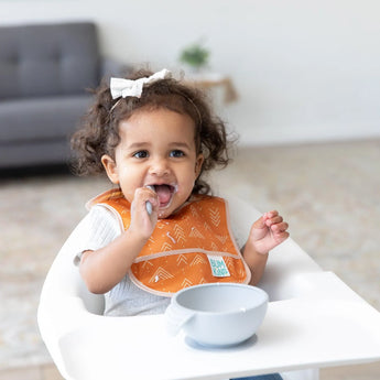 A toddler with curly hair wears Bumkins orange Starter Bib from the Sunshine and Grounded pack, eating with a spoon in a high chair.