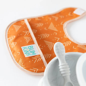 An orange bib from the Sunshine and Grounded set sits beside a white baby bowl and spoon, featuring the Bumkins logo.