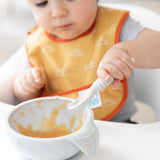 A baby wearing a Bumkins Starter Bib from the Sunshine and Grounded pack is in a high chair with a spoon, eating from a bowl of mushy food.