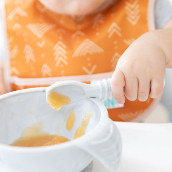 Close-up of a child in a Bumkins Starter Bib, from the Sunshine and Grounded pack, holding a spoon of baby food over a bowl.