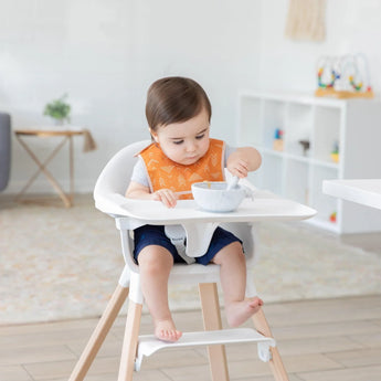 In a modern, bright room, a toddler uses a spoon to eat while wearing Bumkins waterproof Starter Bib in Sunshine orange.