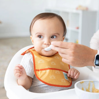 An adult feeds a baby using a spoon while theyre content in their high chair, wearing a Bumkins Sunshine and Grounded Starter Bib.