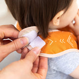 A person fastens a Velcro Starter Bib from Bumkins Sunshine and Grounded pack on a toddler, who holds a finger near their mouth.