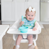 A baby sits in a highchair with Bumkins Starter Bib: Ocean Life, holding a blue spoon over a bowl. The bibs are easy to machine wash.