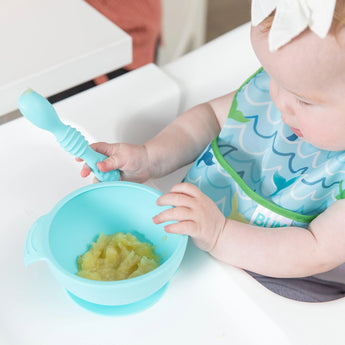 A baby in a washable Bumkins Starter Bib holds a spoon over mashed food on a highchair.