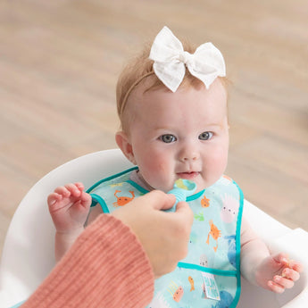 A baby in a blue Bumkins Starter Bib, part of the Ocean Life & Whale Tail set, is spoon-fed, enjoying machine washable ease.