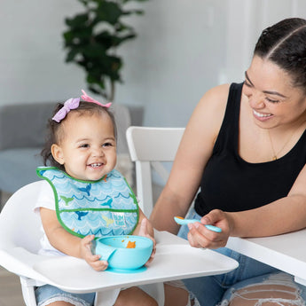 A smiling woman feeds a happy toddler in a high chair using the Bumkins Starter Bib 2 Pack: Ocean Life & Whale Tail with a bowl and spoon.