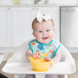 Baby in a high chair, smiling with a Starter Bib by Bumkins in Ocean Life print, holding a yellow bowl and spoon.