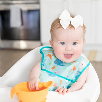 Baby with a white bow wears Bumkins Ocean Life Starter Bib, sits in a high chair holding a spoon in an orange bowl, smiling.