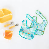 Two vibrant Bumkins Starter Bibs, plus yellow plate with utensils and cup—all crafted from machine washable, waterproof fabric.