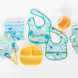 Bumkins Starter Bib 2 Pack: Ocean Life & Whale Tail with a yellow plate, orange cup, utensils, and sandwich bags.