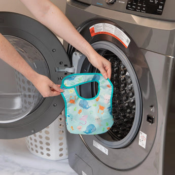 Someone washes a Bumkins Starter Bib 2 Pack: Ocean Life & Whale Tail, with waterproof fabric, in a front-loading washing machine.