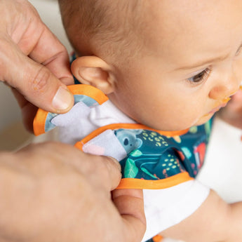 An adult fastens a Bumkins Starter Bib with orange trim from the Jungle & Animal Prints pack on a baby, who gazes to the side.