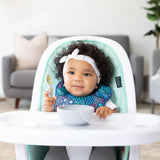 A baby in a high chair wears a Bumkins Starter Bib from the Jungle & Animal Prints 2 Pack, holding a spoon and eyeing the bowl.