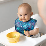 A baby in a high chair wears a Bumkins Starter Bib with jungle print, smiling and holding a spoon while eating from a yellow bowl.