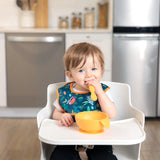 A toddler in a high chair, wearing a Bumkins Starter Bib from the Jungle & Animal Prints pack, holds a spoon and sits by a bowl.