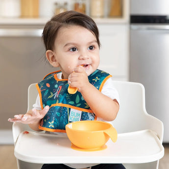 A baby in a Bumkins Starter Bib with animal prints is in a high chair, holding a spoon, eating from an orange bowl.