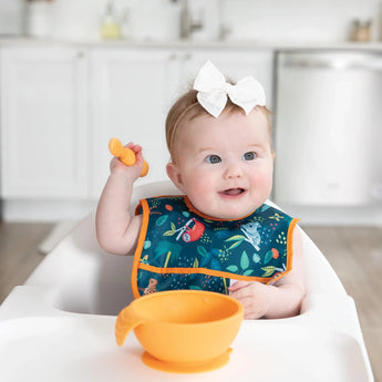 A baby with a white bow sits in a high chair, holding an orange utensil. She wears a vibrant Bumkins Starter Bib from the Jungle & Animal Prints pack made of safety-tested, waterproof fabric. An orange bowl is set on the tray before her against a kitchen backdrop with white cabinets.