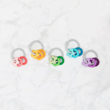 Five Silicone Teething Charms: Orange by Bumkins sit on a white marble surface, featuring interlocking gender symbols.