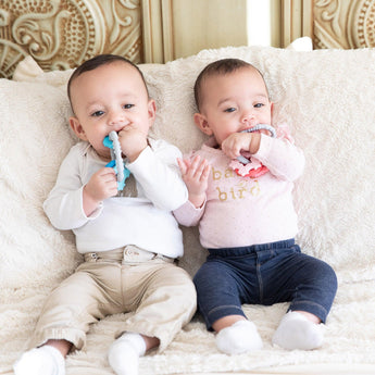 Two babies on a couch with Bumkins Silicone Teething Charms: Blue. One in a white shirt, the other wears pink with baby on it.