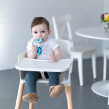 A baby in a high chair chews on Bumkins Silicone Teething Charms: Blue, enhancing motor skills, in a bright room with flowers.