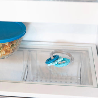 Inside the fridge, a blue-lidded glass container is next to Bumkins Silicone Teething Charms: Blue.