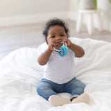 A baby, wearing a white shirt and jeans, chews on Bumkins Silicone Teething Charms: Blue, sitting on a white blanket.