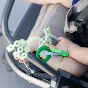 Baby holds a green Silicone Teething Charm by Bumkins, ideal for developing motor skills in a stroller. Made from food-safe silicone.