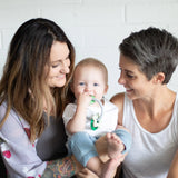 Two women smile at a baby with Bumkins Silicone Teething Charms: Green, happily seated on ones lap against a white wall.