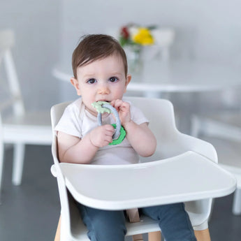 A baby in a high chair chews on Bumkins Silicone Teething Charms: Green, with a blurred table and flowers in the background.