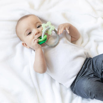 Baby lying on a white blanket, grasping Bumkins green Silicone Teething Charm, wearing a white shirt and gray pants.