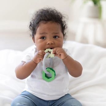 A baby in a white shirt and jeans sits on a blanket, chewing on a Bumkins Silicone Teething Charm: Green to develop motor skills.