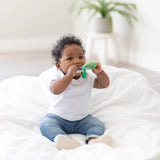 A curly-haired toddler sits on a white blanket, wearing a white shirt and blue jeans, while chewing on Bumkins Silicone Teething Charms in green for teething relief. A blurred potted plant in the background enhances the calm, cozy setting.